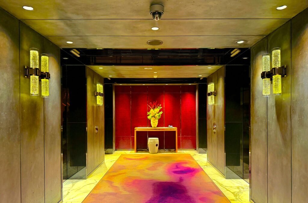 Luxurious hallway with vibrant pink and orange rug, glowing wall lights, and a table topped with a colorful flower arrangement against a rich red backdrop.