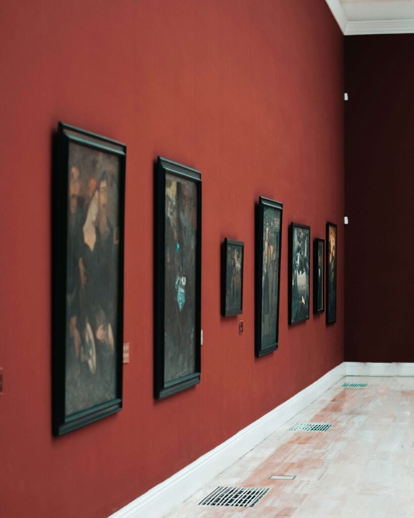 Paintings in black frames line a deep red gallery wall, creating a classic and elegant atmosphere. The light wood floor adds warmth and balance.