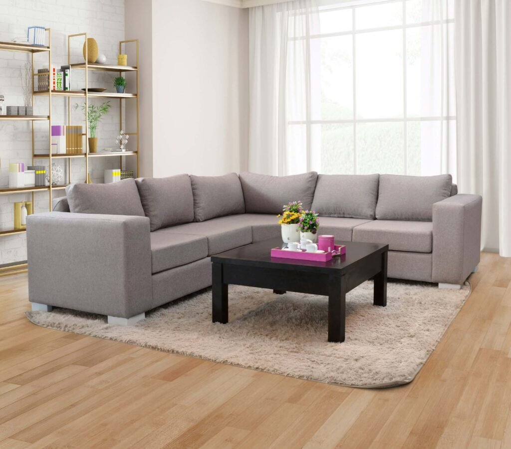 Modern living room with a gray sectional sofa on a beige rug, facing a dark coffee table. Bookshelves and large window enhance the bright, cozy ambiance.