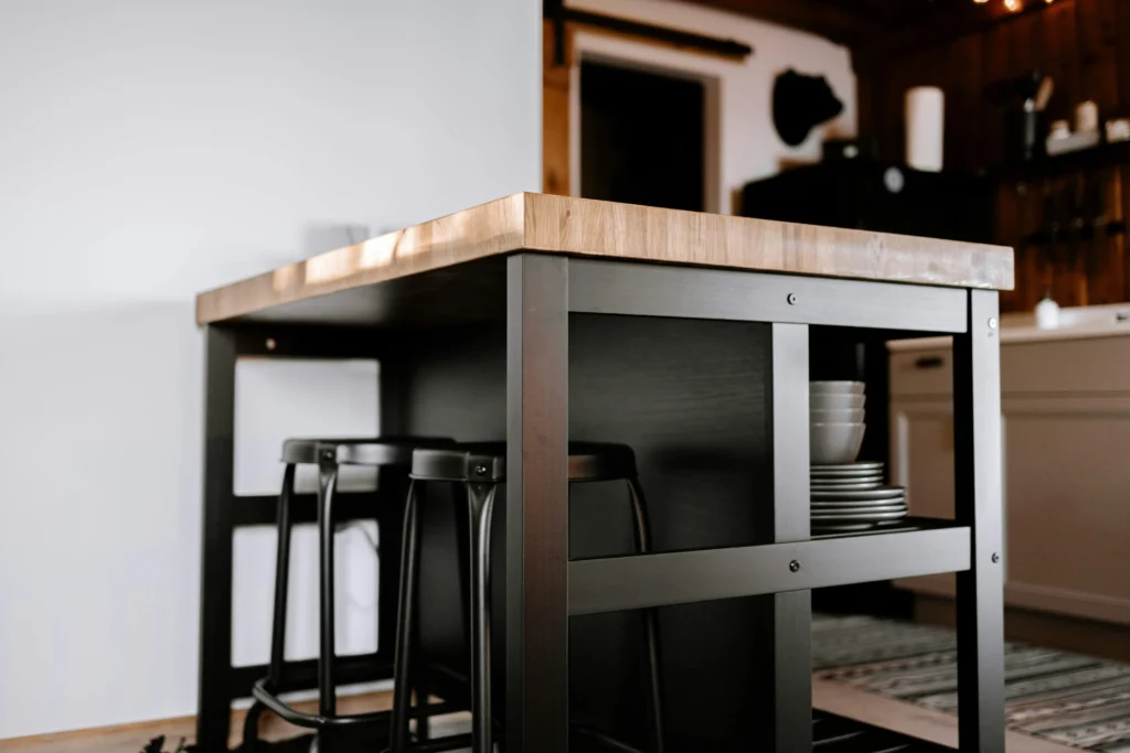 A modern kitchen island with a wooden top and black metal legs, featuring two stools and stacked white dishes. The setting is warm and inviting.