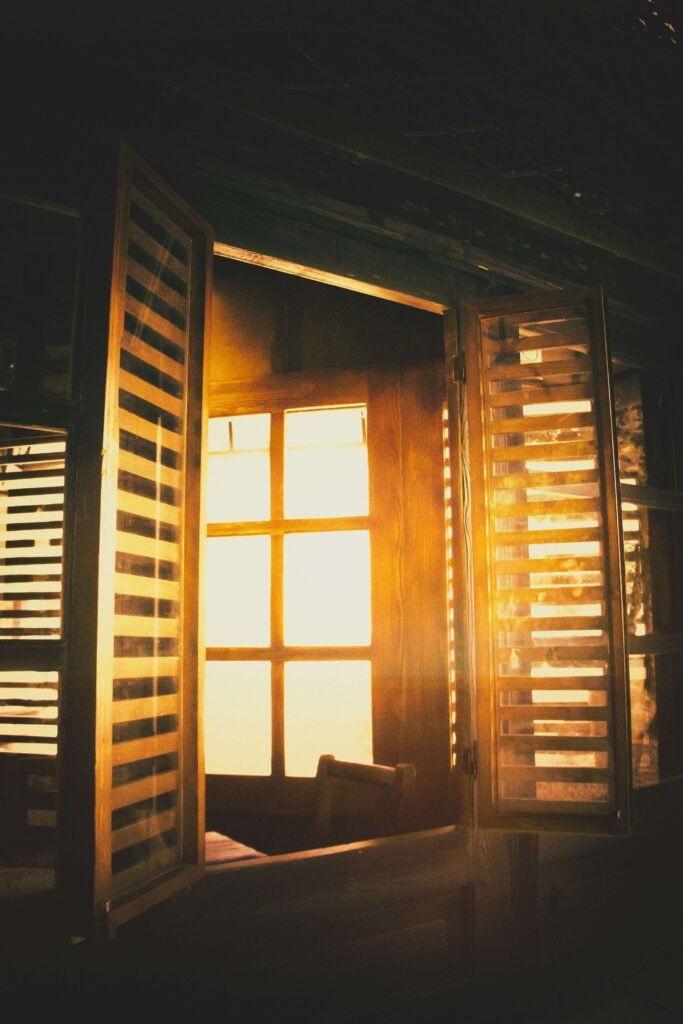 Warm sunlight streams through open wooden shutters of a window, casting soft, golden light and shadows inside, creating a cozy, nostalgic atmosphere.