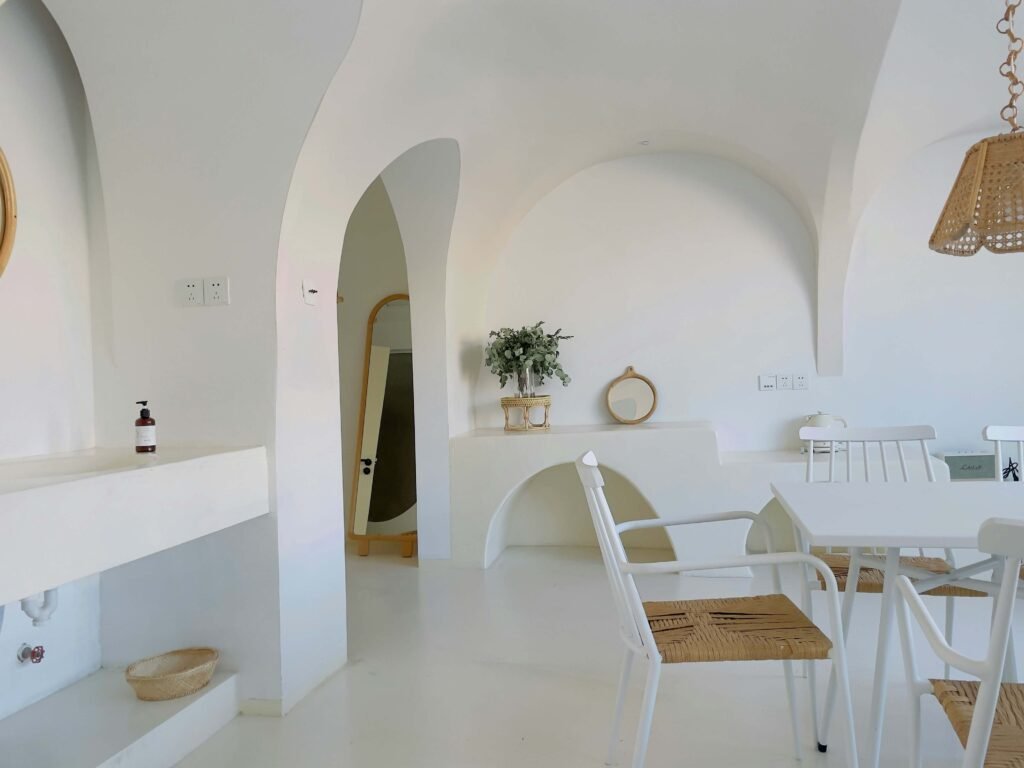 A minimalist white room with arched doorways and walls, featuring a white table with wicker chairs. Greenery and decor add a touch of nature and warmth.