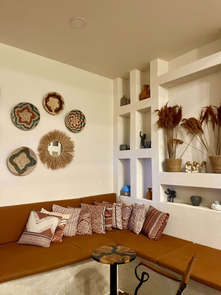 Cozy corner with a brown couch adorned with patterned pillows, geometric wall art, wicker baskets, and shelves with vases and pampas grass. Earthy tones.