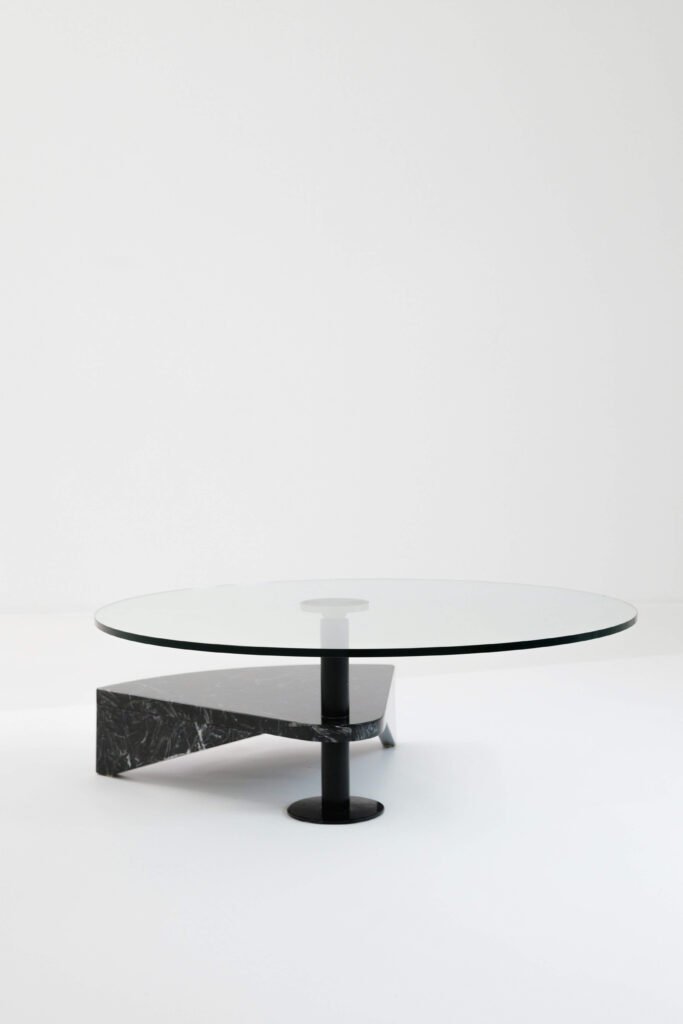 A minimalist table with a round glass top supported by a sleek black metal pole and an angular black marble base, set on a plain white background.